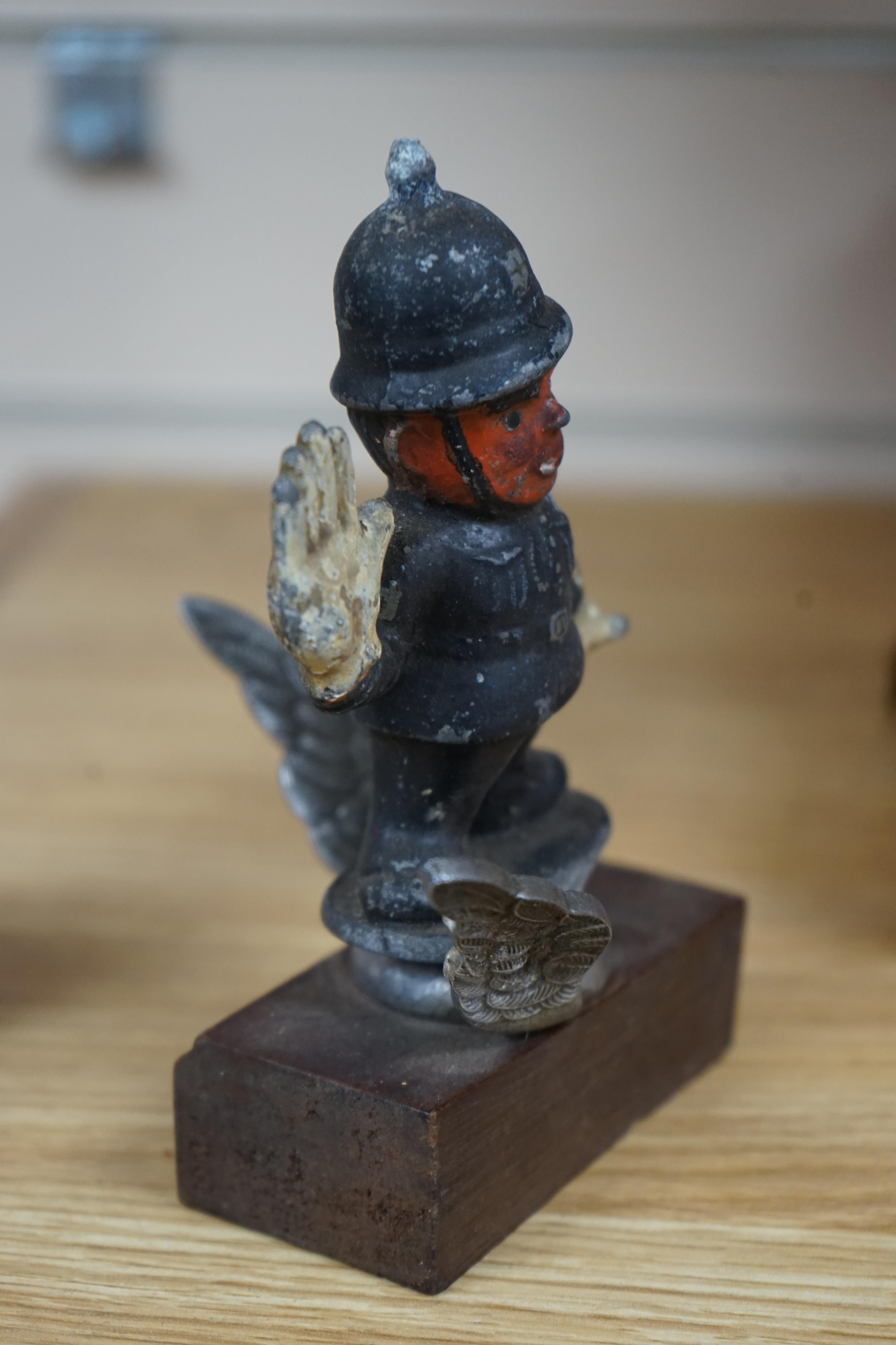 A cold painted cast aluminium car mascot in the form of a caricature of a policeman on associated chromed wings, mounted on a wooden block, policeman 11cm high. Condition - poor to fair.